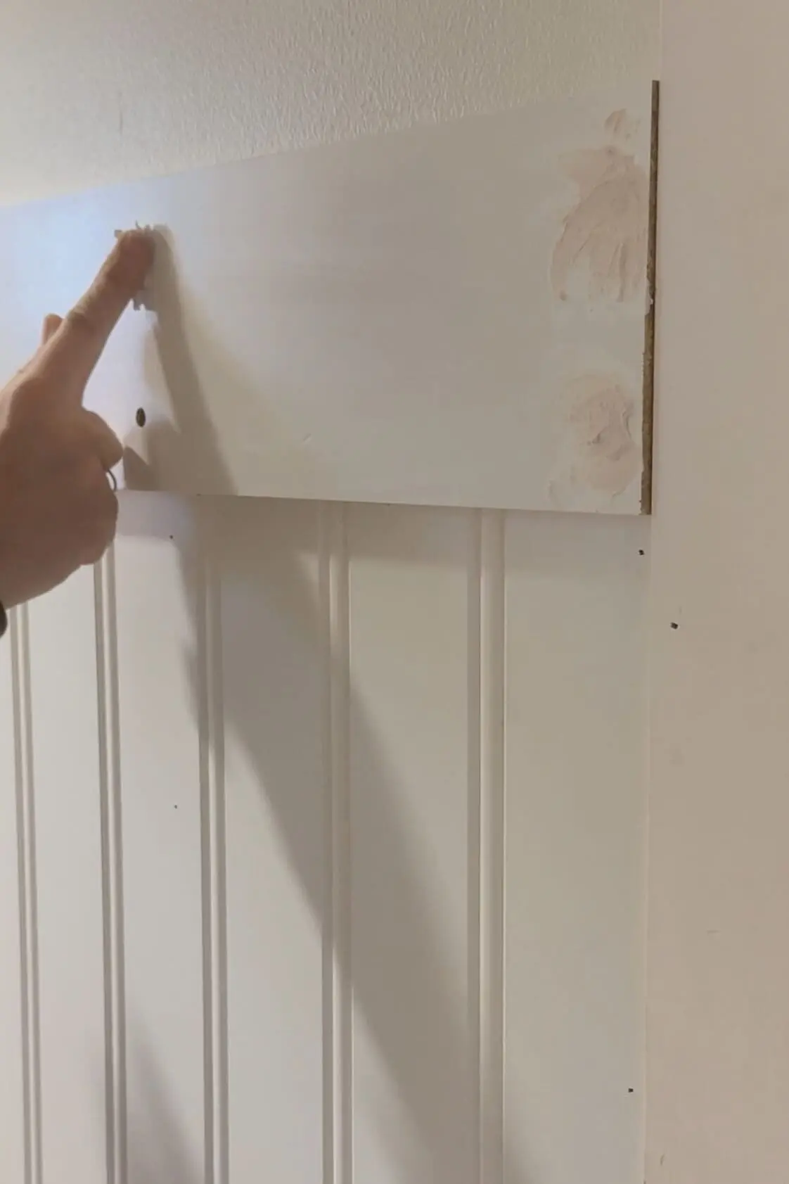 A close-up of a woman’s hand applying spackle to screw holes on a wooden peg rail mounted on beadboard paneling. The smooth wall surface and filled gaps reflect progress in the DIY project.